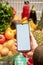 hand holding phone with white screen copy space in grocery shopping cart with fruits and vegetables