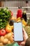 hand holding phone with white screen copy space in grocery shopping cart with fruits and vegetables