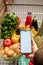 hand holding phone with white screen copy space in grocery shopping cart with fruits and vegetables