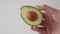 Hand holding a sliced half of a fresh juicy avocado with a bone on a white background.