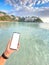 Hand holding a smartphone on the turquoise beach with mountains in the background, unrecognizable person, blank mobile screen