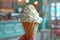 Hand holding vanilla ice cream scoop in a waffle cone against parlor background. Generative AI