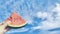 Hand holding a watermelon, blue sky and white clouds background.