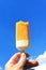 A hand holds a bitten-off ice cream in yellow icing against a blue sky. Refreshing summer dessert.