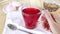 Hand holds cup of fruit tea and turns it on table. Hibiscus in glass mug. Bright red hot drink, compote. Summer tea break with