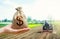 A hand holds out a dollar money bag on a background of a field cultivating tractor. Lending farmers for purchase land and seed