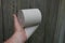 A hand holds a roll of gray toilet paper against a wooden wall