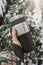 A hand i holding a Reusable Thermo mug, thermos, travel mug on the background of a snow-covered coniferous tree, winter outside.