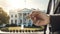 the hand of a man in a suit holds a key against the backdrop of the White House