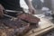 Hand with metal clamp grabbing grilled meat on a cutting wood, blurred person in the background.