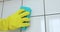 Hand of person in yellow glove is washing white tile using blue sponge.