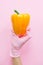 Hand in pink glove holding yellow pepper on pink background flat lay. Order groceries and get them delivered safe during