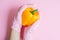 Hand in pink glove holding yellow pepper on pink background flat lay. Order groceries and get them delivered safe during