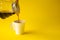 Hand pouring liquid coffee put in the cup on a yellow paper background. Copy space for your text