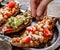 Hand pouring sesame seeds on baked sweet potato toasts with roasted chickpeas, tomatoes, goat cheese, avocado, seedlings
