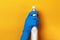 A hand in a protective latex glove holds a spray bottle on a yellow colored background