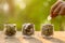 Hand putting coins into clear money jar on wooden table with green blur light background. Savings concept