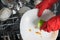 A hand in a red protective glove, cleans white plates of large food debris, for putting in the dishwasher, close-up