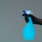 A hand in a rubber glove holds the glass cleaner in a spray bottle on a neutral background.