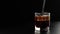Hand stirs ice cubes with cola using a black straw. Ice floats with bubbles in a glass of soda with brown highlights