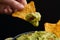 Hand with tasty tortilla chips or nachos with fresh homemade guacamole dip sauce on black background