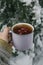 A hand in a warm sweater holding mug cup with hot healthy herbal tea with dog-rose rosehips on the background of a snow-covered co