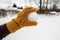 A hand in a yellow mitten holds a snowball, in the yard against the background of a snow-covered vegetable garden