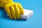 Hand in yellow protective glove with white melamine household sponge on blue background. Means for cleaning surfaces