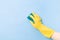 A hand in a yellow rubber glove holds a yellow sponge with a green rough side for cleaning complex dirt, blue background