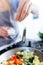 Hand of a young woman adding salt to vegetables into the pan.