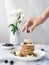 The hand of a young woman decorates with blueberries a stack of pancakes.