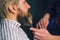 Hands of barber making haircut to attractive bearded man in barbershop.