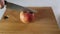 A hands cuts a red apple with a knife on a wooden board.
