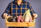 Hands of a farmer holding a wooden box with avocados, purple yams, and purple corn while standing on a gray background.