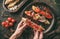 Hands holding a bruschetta with prosciutto, tomatoes, parmesan cheese, sauce pesto, grilled pepper on ciabatta bread on wooden