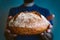 Hands holding rustic and crusty round loaf of fresh whole grain bread on dark blue background with white flour on top. making