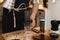 Hands holding steel kettle and aeropress, glass cup, scales, grinder on wooden table. Professional barista pouring hot water in