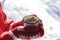 Hands in Knitted Mittens holding Steaming Cup of Hot Tea on Snowy Winter Morning Outdoors. Woman holds Cozy Festive Red