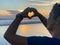 Hands making love sign. Fingers with heart gesture. Sunset in hand on blue beach landscape background. Love hope faith concept.