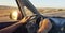 Hands of a man driver on car steering wheel. Dashboard. Windshield and side window view. Arid countryside landscape along the road