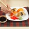 Hands taking ginger with chopsticks. Assorted sushi and soy sauce on black and red bamboo mat.