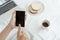 Hands of working woman holding and using smart phone on desk background. On the desk have cup coffee, breads and laptop