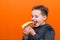 Handsome 10 yers old boy holding and biting hot dog closeup indoors orange studio background image.Close up,copy space