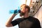 Handsome adult man drinking water from fitness bottle while standing outside, at sunset or sunrise. Runner.