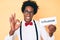 Handsome african american man with afro hair holding paper with influencer text doing ok sign with fingers, smiling friendly