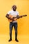 Handsome african american retro styled guitarist playing acoustic guitar isolated on yellow background.