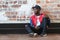 Handsome African man in casual clothes and a cap is sitting on the floor in slum on background of ruined red brick wall