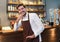 Handsome bearded man in apron looking at camera and holding hand on chin while standing at front bar counter. Startup small