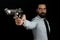 A handsome bearded man on black background holds a gun in his hand pointing it in front of him.