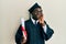 Handsome black man wearing graduation cap and ceremony robe holding diploma serious face thinking about question with hand on